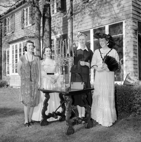 Four women attending the Republican Centennial Birthday party are shown modeling in the historical fashion show. Pictured from left to right are: Mrs. Charles Vaughn, in a 1924 flapper dress; Mrs. Wade Plater, in a 1860 costume; Mrs. Stewart Honeck, in a present day 1954 suit; and Carol Rennebohm in a 1904 dress and hat, holding a fan. They are all watching Mrs. Honek light the candles on a candelabra.