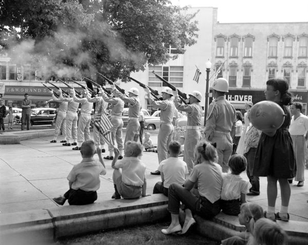 Firing Squad of the 520th Air Defense Group at the Memorial Day parade around Capitol Square. The servicemen are aiming their rifles in the air as children are looking on. One girl is holding a balloon, and another has her fingers over her ears. 