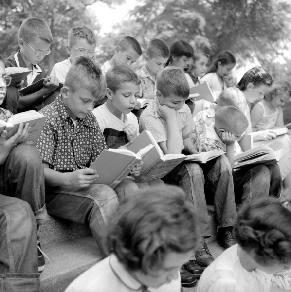 The book, <i>Days of Adventure</i>, being read aloud by Hadi Pankow and Randy Sumner as other boys and girls follow along. The group is sitting on stone steps.