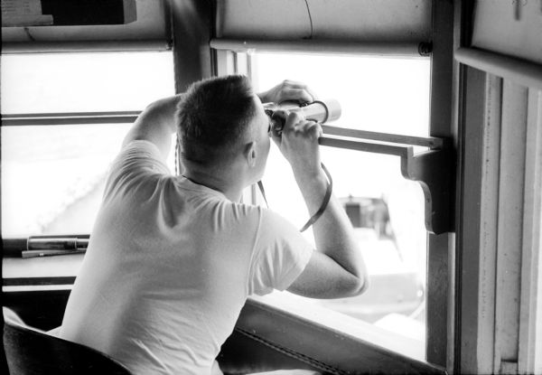 Robert Risser, Jr., of 3806 Hammersley Road, on duty watching for boaters in distress on Lake Mendota from the U.W. Lifesaving Services lookout. He is using binoculars. The lifesaving services facility located in the old university boathouse was originally at the current site of the U.W. Alumni Building behind the Red Gym.