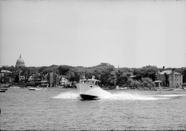 View across water towards the U.W. Lifesaving Services rescue boat, "Isabel III," with Captain Harvey Black at the helm speeding to assist a distressed boater on Lake Mendota. The Capitol dome is visible in the distance, beyond the lakeshore. The lifesaving services facility located in the old university boathouse was originally at the current site of U.W. Alumni Building behind the Red Gym.