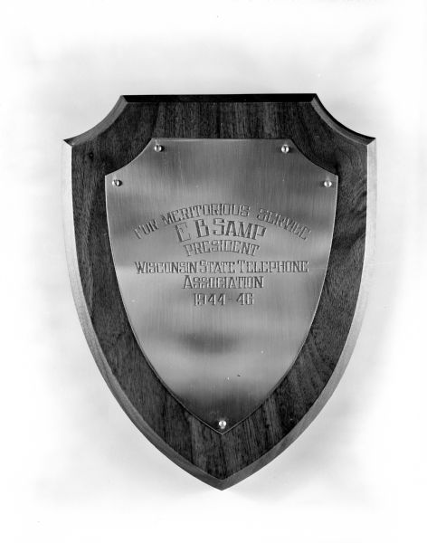 A brass plaque shaped like a shield with the following inscription: "For Meritorious Service - E. B. Samp President - Wisconsin State Telephone Association - 1944-1946."