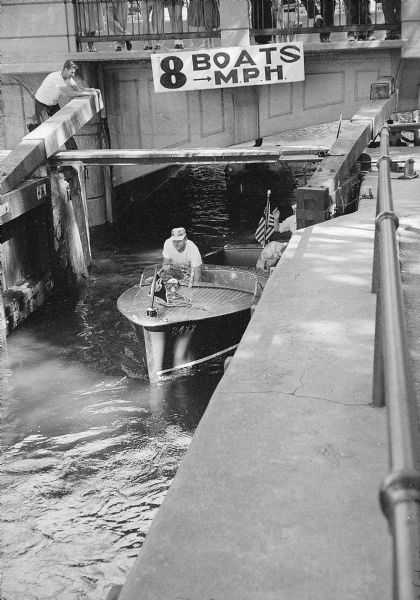 Locktender Earl Wood swinging open the heavy lock gate for a speedboat operator passing through the lock on the Yahara River at Tenney Park. A sign above the lock reads: "Boats - 8 MPH."
