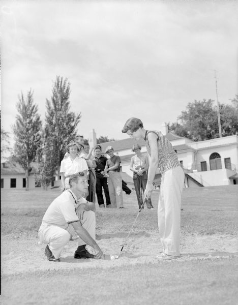Man squatting down pointing to a golf ball in a sandtrap at Maple Bluff Country Club, instructing a woman to how to hit it. There are several onlookers behind them on the green. The club house is in the background. 