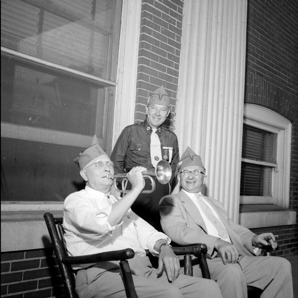 C.C. Benn is shown warming up his bugle. He blew taps at the Wisconsin American Legion's 40 and 8 memorial service. Looking on are Vig Iversen and Jacob Mintz. They are on the porch of the Elks Club.