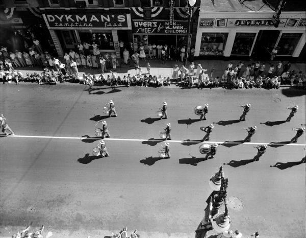 Elevated view looking down at a marching band participating in the Wisconsin Department of the American Legion annual convention parade around the Capitol Square. The marchers' shadows are preceding them as they march past Dykman's Tempting Food Store and Dyers' Shoes for Men, Women, and Children.