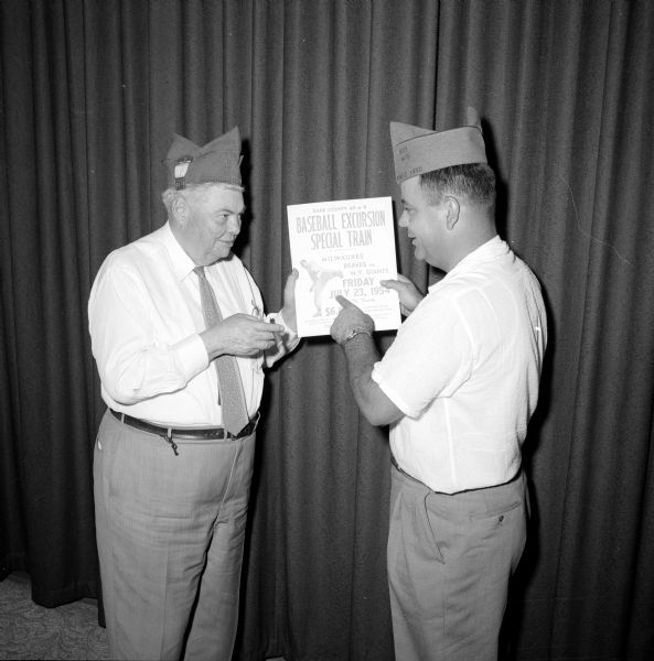 S.C. Hull, Milton Junction (left), and Bill McCunnell (of McFarland) are holding a poster promoting the Dane County American Legion 40 and 8 baseball excursion to a Milwaukee Brewers baseball game. The men were attending the state 40 and 8 convention at the Elks Club held prior to the American Legion convention at the Hotel Loraine.
