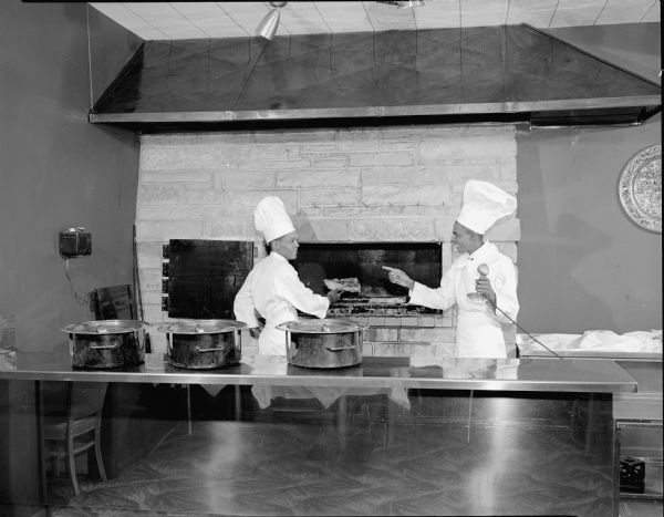 Two African American cooks standing by a stone hearth in the kitchen at the New Pines restaurant located at 2413 Parmenter Street.  One of the cooks might be "Chef Joseph" — he is mentioned in an ad for the restaurant in June 1954.
