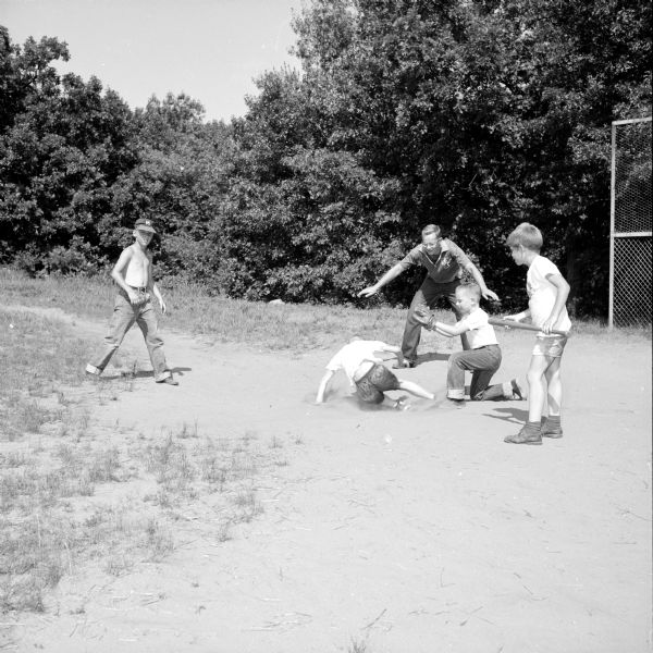 YMCA staff member Harold Haak (top, center) declares Bill Cunningham (left) safe at home. Other players are Robert Berhagen as catcher and Steve Walrath, waiting to bat. The boy on the left is unidentified.