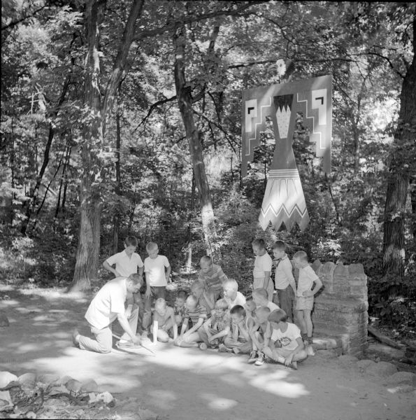 YMCA Camp superintendent Al Bagley (left) explaining a project to eighteen boys at the camp council ring. Behind them a large eagle cutout is hanging from a tree branch, and it is painted with American Indian motifs. 