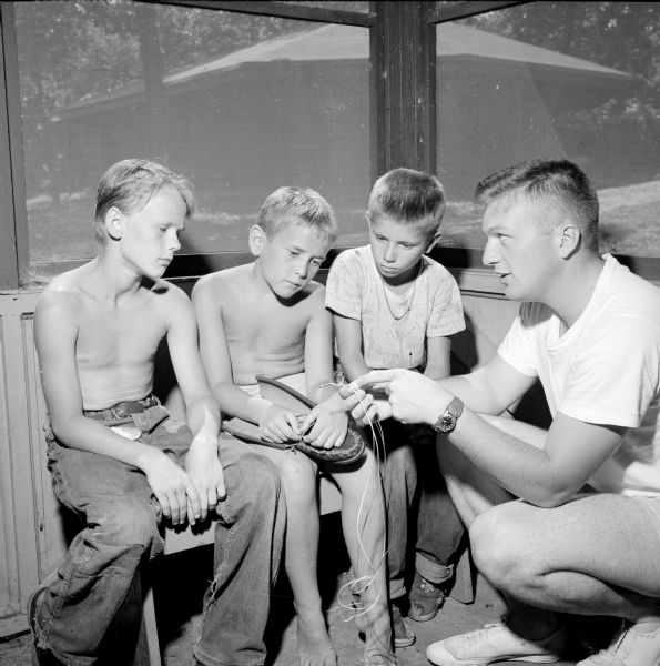 Three boys receiving craft instruction in how to braid thongs for use in making a pair of "Indian" moccasins from Dick Carman (right). The boys (left to right) are Daniel Steinhoter, George Schroeder and Dale Weitzel.