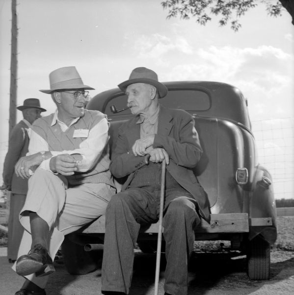 Early settler Anton Thomas, sitting on the right on the wooden bumper of an automobile, is holding his cane and smoking a pipe, and is talking with Robert C. Birkinbine sitting on the left.