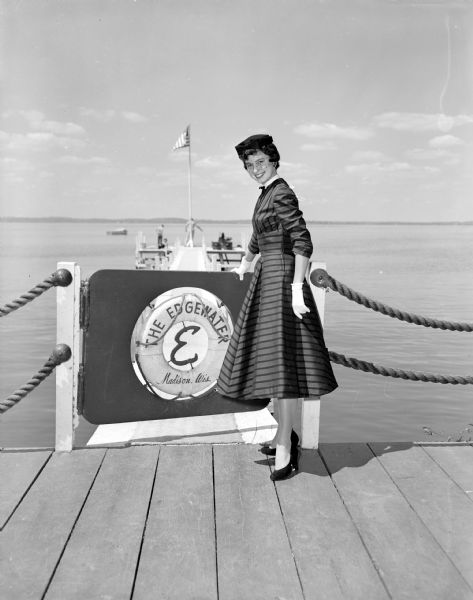 Fashion photo for Woldenberg's taken on the Edgewater Hotel Pier located on Lake Mendota at the end of Wisconsin Avenue. A woman in a striped dress, dark hat, and white gloves is posing with her hand on a gate that leads to the pier. Behind her, a United States flag is flying at the top of a pole at the end of the pier.