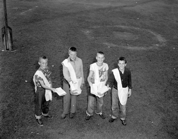 Elevated view of four newspaper boys for the <i>Wisconsin State Journal</i>. Looking up at the photographer and holding out folded papers, they are wearing cuffed blue jeans and wearing their satchels over their shoulders.