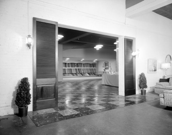 Entrance to the newly enlarged floor covering and drapery display area at Black Furniture Company, located at 315 West Gorham at State Street. Carpet samples are on a back wall, seen from a doorway in another display room, which is decorated with sconces and small evergreen trees in planters.