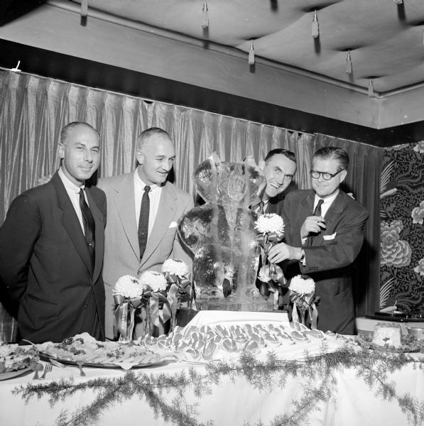 Four men standing at a banquet table with a Bucky Badger ice sculpture as the centerpiece. One is holding one of the chrysanthemums that decorated the table by Bucky's paw.