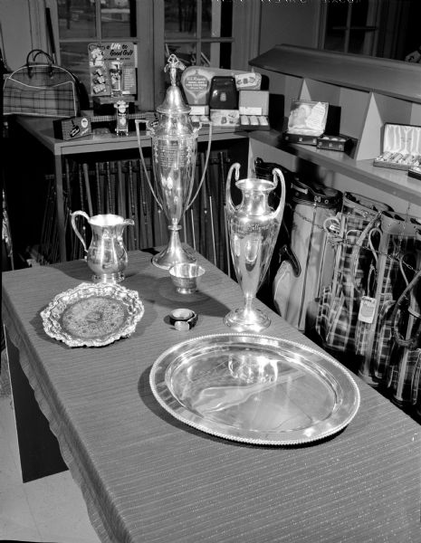 Table displaying Maple Bluff Country Club trophies and commemoration plates in the Pro Shop. Behind the table are counters with small golf items for sale, and underneath are golf clubs and golf bags.