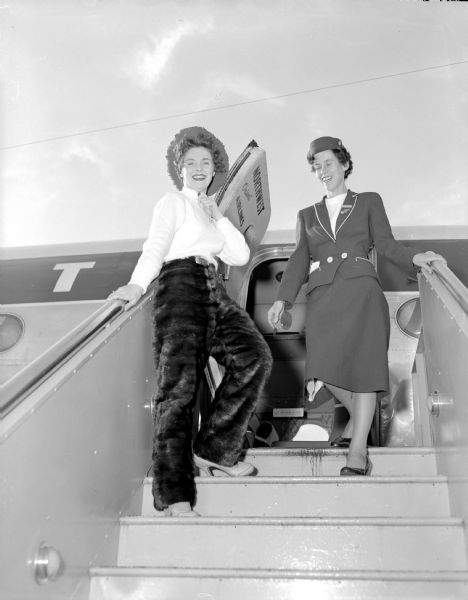 A model, and a North Central Airlines stewardess posing at the top of loading stairs leading up to an airliner for Northwest Airlines. The model is posing in mink pants, a cowboy hat, and high heeled shoes. The shoot was for the Emporium Department Store.