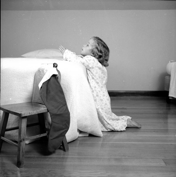A young girl kneeling at the side of a bed accompanies a plea for donating to the Empty Stocking Club organized by the <i>Wisconsin State Journal</i>. A stocking is hanging in the foreground over the back of a chair.