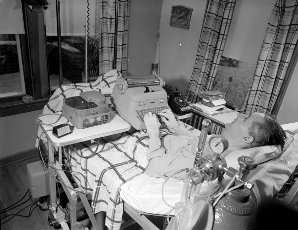 A bed-ridden man working from home. Laying in a hospital bed set up in his living room, he is using a typewriter that that is sitting on a table between a rotary-dial telephone on one side, and a record player and intercom on the other. An oxygen tank and tubes are by the head of the bed in the foreground. His name and address are unknown.
