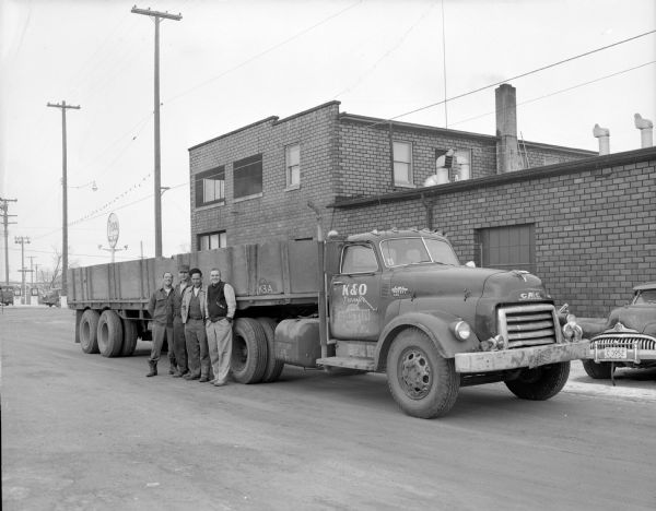 Four men standing in front of a K & O Transfer truck pulling a long flatbed at an unknown location. They are right behind a building and down a back alley from a gas station.