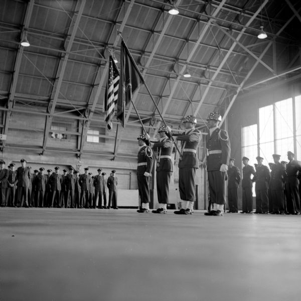A Truax Field color guard stands at attention, and Truax Field personnel are at parade rest in a field hangar at the start of a ceremony honoring Chief Warrant Officer Sam Grubner, who retired after more than 30 years in the Army and Air Force.
