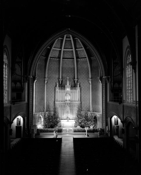 Night time elevated interior view of Grace Episcopal Church, located at 116 West Washington Avenue. Taken from an elevated vantage point in the balcony, the photograph centers on the high altar at back in the sanctuary across the darkened nave. Stained glass windows are on the side walls. The altar is decorated with pine trees for Christmas.