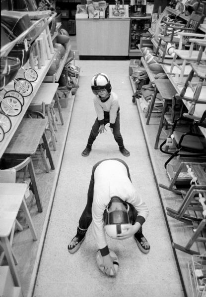 Wearing helmets, Jeff and Gregg Anderson playing with a football in the Wolff-Kubly-Hirsig store, Toyland. They are in an aisle displaying rocking chairs, small tables and chairs, and baby strollers.