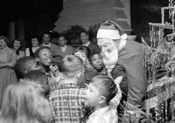 Al Sitniek, wearing a Santa Claus mask and costume, is crowded by black and white children at a Christmas party. The event was organized for children of the South Side Community Center by Delta Tau Delta Fraternity at 16 Mendota Court.