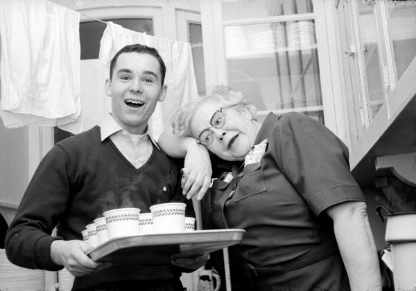 Fraternity member, James Slone, smiling and holding a tray of hot beverages while his fraternity's housemother, Mrs. Dorothy Lambert, is leaning her arm and head on his shoulder. They are hosting a party for children of the South Side Community Center at the fraternity on Mendota Court.