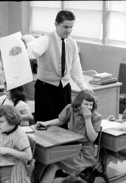 Art instructor Ray Lenahan instructing a third-grade class at Nichols School in the art of finger painting using an old-style Crayon drawing as an example, while student Julie Stoll (of 5400 Harold Avenue) is sharing a laugh with another student.