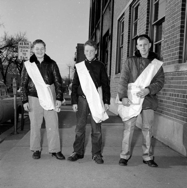 Three <i>Wisconsin State Journal</i> newspaper boys. They are standing on a city sidewalk by parking meters with satchels slung over their shoulders and papers in their hands.