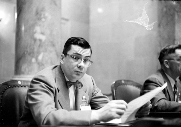 Assemblyman Carroll Metzner (R-West Madison) is listening to a presenter at a pre-session lawmaker's school for the freshman members of the 1955 Wisconsin State Legislature. He is wearing a name tag in the shape of the State Capitol building on his lapel. 