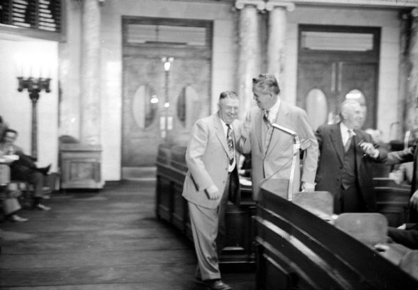 1955 Wisconsin State Assembly members William Loy (R-Landcaster) on the left and Michael F. O'Connell (D-Milwaukee) on the right watching as Speaker-elect Mark Catlin, Jr. (center) is walking to the Assembly Chair position for his first time.