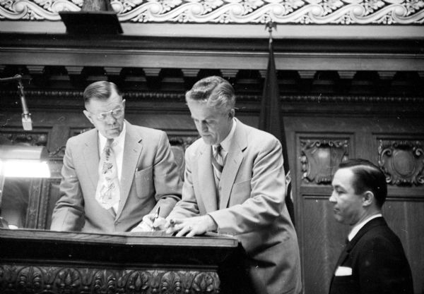 The newly elected Assembly speaker, Mark Catlin, Jr. (right), signing the official vote register as Robert H. Boyson (left), voting machine operator, is looking on. The third man on the far right is unidentified.