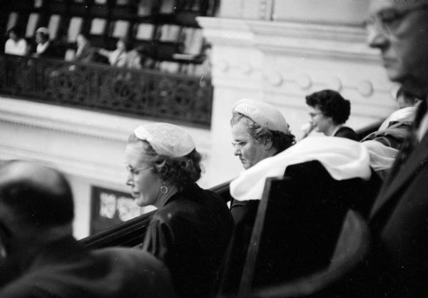 Proud assemblymen's wives watching from the state assembly gallery as the 1955 State Assembly session begins. Mrs. Earl Warren (sitting on the left) is married to the Democrat representing Racine, and Mrs. Margaret Lourigan (right) is married to Joseph Lourigan (D-Kenosha). The woman on the far right without a hat is unidentified.
