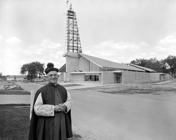 Reverend Monsignor Francis Leo McDonnell, pastor, poses in front of the new Our Lady Queen of Peace Catholic Church located at 401 South Owen Drive. The steeple is seen under construction. 