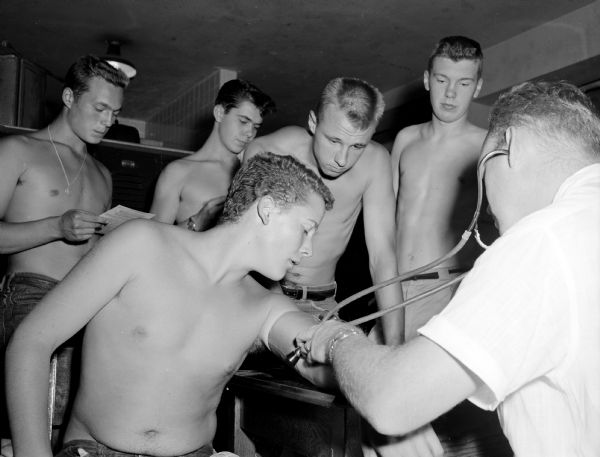Madison West High School football player, Dave Koepcke (seated), is getting his blood pressure checked by Dr. William Walter, who is using a stethoscope during the high school athletes' physicals. Standing in back are Tom Whitmore, Ted Neesvig, Leroy Abrahamson and Ron Anderson. The five athletes are shirtless, and all have short hair.