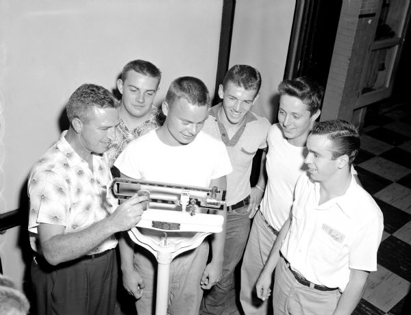 Coach Harold (Gus) Pollock (left) adjusting the counter weights on the scale being used by Ron Morrick. Other returning veteran players shown are Steve Schleck, Bernard Kane, Dennis Schmelzkopf, and Bill Connors.   