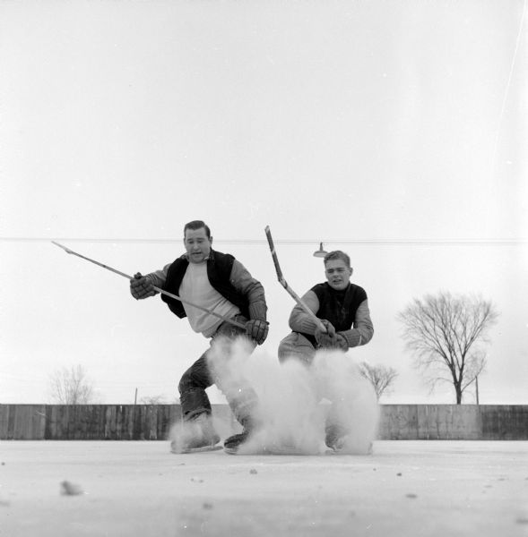 Bob Lake and Dick Wills, holding their sticks up, stopping abruptly in pursuit of a hockey puck at Vilas park rink.