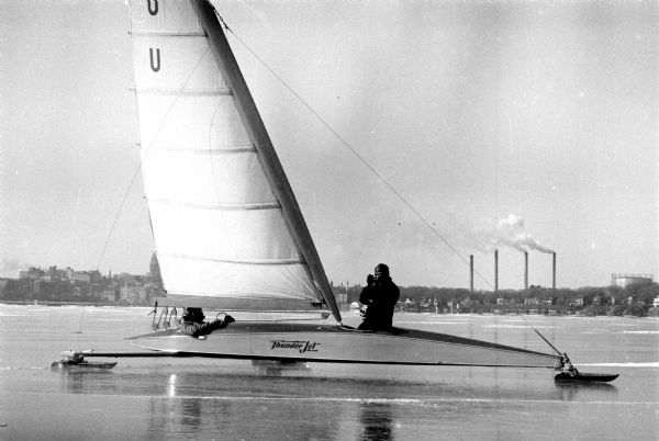 The ice boat, "Thunder Jet," on a frozen Lake Monona. One of the two men aboard is probably the pilot, Bill "Curley" Perrigo, sitting at the helm or by the mast. Madison Gas and Electric exhaust stacks are on the horizon, and the Wisconsin State Capitol can be partially seen behind the sail.  