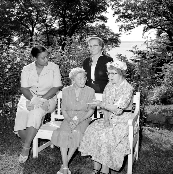 Four women sitting around a bench in a garden by Lake Mendota. They are discussing plans for the Annual World Fellowship Benefit Garden Tea sponsored by the City YWCA. They are (left to right): Mrs. William Jenkins in white gloves, Mary Hanks, Violet Kingsbury, and Caroline Kirkpatrick.