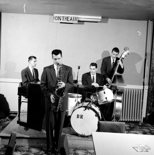 Four men playing musical instruments in a studio with a electric sign that says: "On the Air." U.W. student Ray Price led the band, which consisted of stand-up base, drums, piano and saxophone.