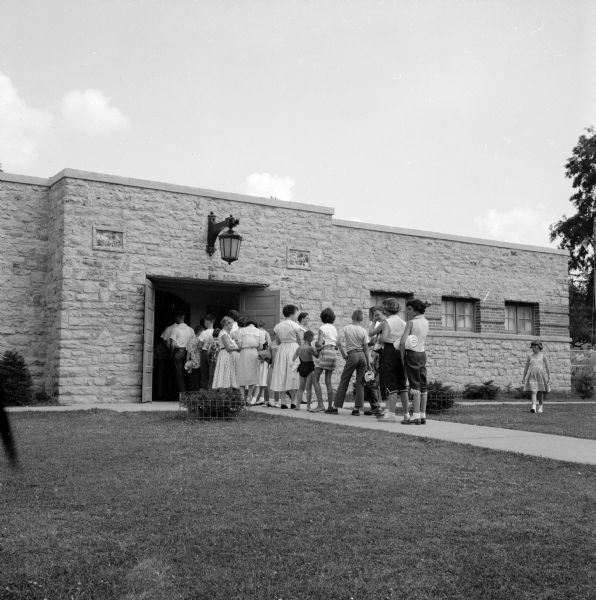 Children lining up on a sidewalk to enter the stone bathhouse that leads to the swimming pool.