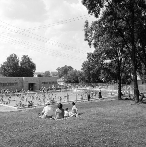 Parents and children sitting on a blanket on a lawn and looking down over the swimming pool, which is enclosed by a chain-link fence at Badger Park.