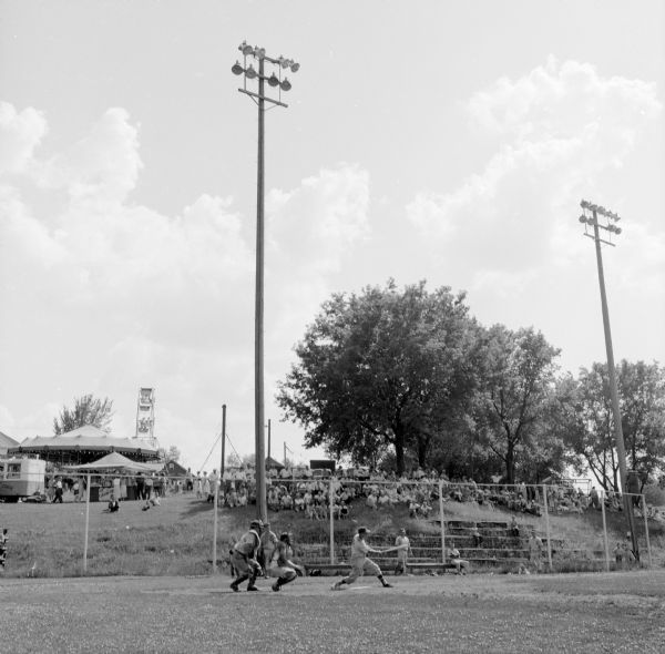 Norman Schandt, a member of the Shullsburg team, batting against the Potosi team in a baseball game at Badger Park. A crowd of spectators outside the fence are sitting under the shade.
