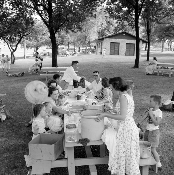 A family picnic at Badger Park includes the Mulcahy, Solberg, O'Neil and Foley families. All of the families are from Shullsburg. One mother is placing a large crock pot at one end of the picnic table and a father is unpacking items at the other. A couple children have balloons.