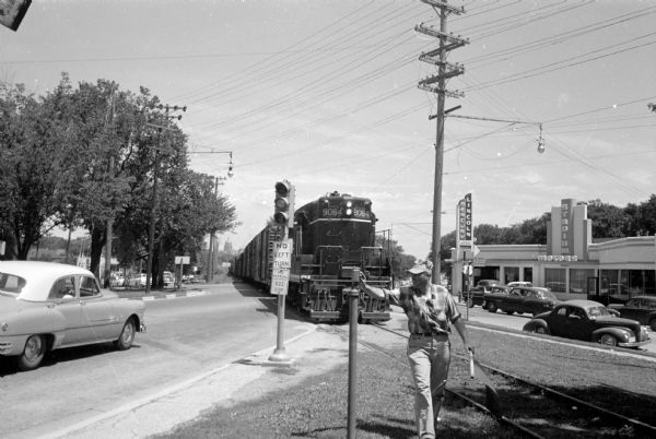 Brakeman R.D. DeRoche pressing the button to deactivate the warning lights as the Illinois Central train passes the intersection with Monroe and Regent Streets. The Lincoln Mercury Stadium Motors car dealership is in the background.