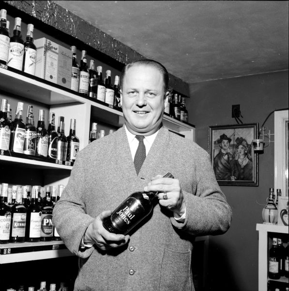 Man holding a bottle of Paul Jones liquor in front of other brands on a wall shelf.