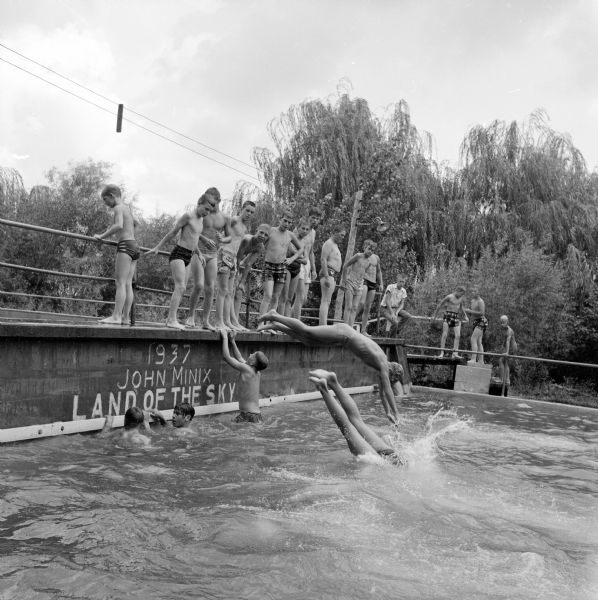 Nineteen <i>Wisconsin State Journal</i> carrier boys at the Blue Mounds State Park swimming pool. Some are shown diving off a platform named for the previous park owner, John Minix, and dated 1937 with the slogan "Land of the Sky." The boys came on four charted buses for a picnic hosted by the journal.